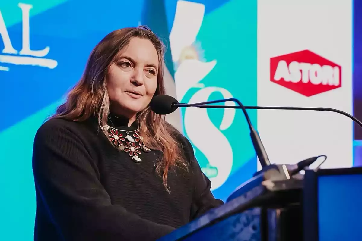 Mujer hablando frente a un micrófono en un escenario con fondo azul y logotipos de empresas