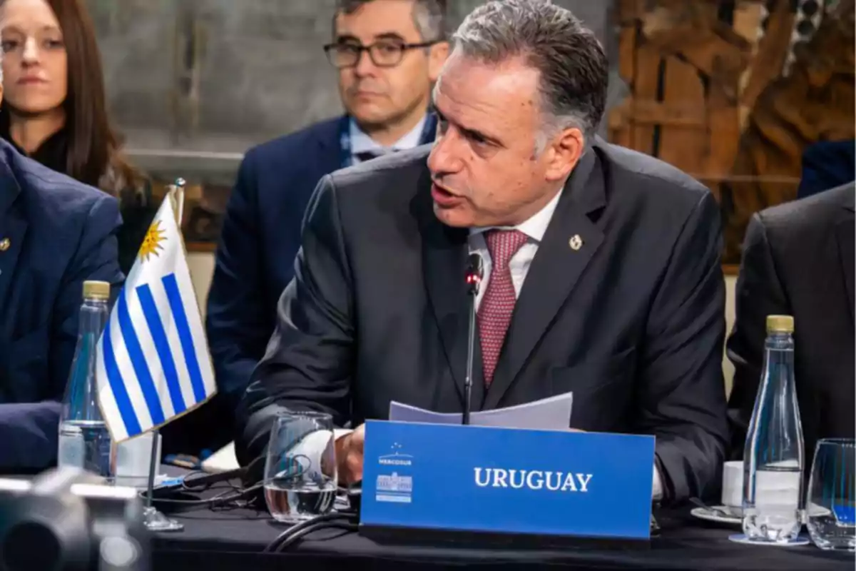 Man in a suit speaking at an international conference with a Uruguay sign and a Uruguayan flag on the table
