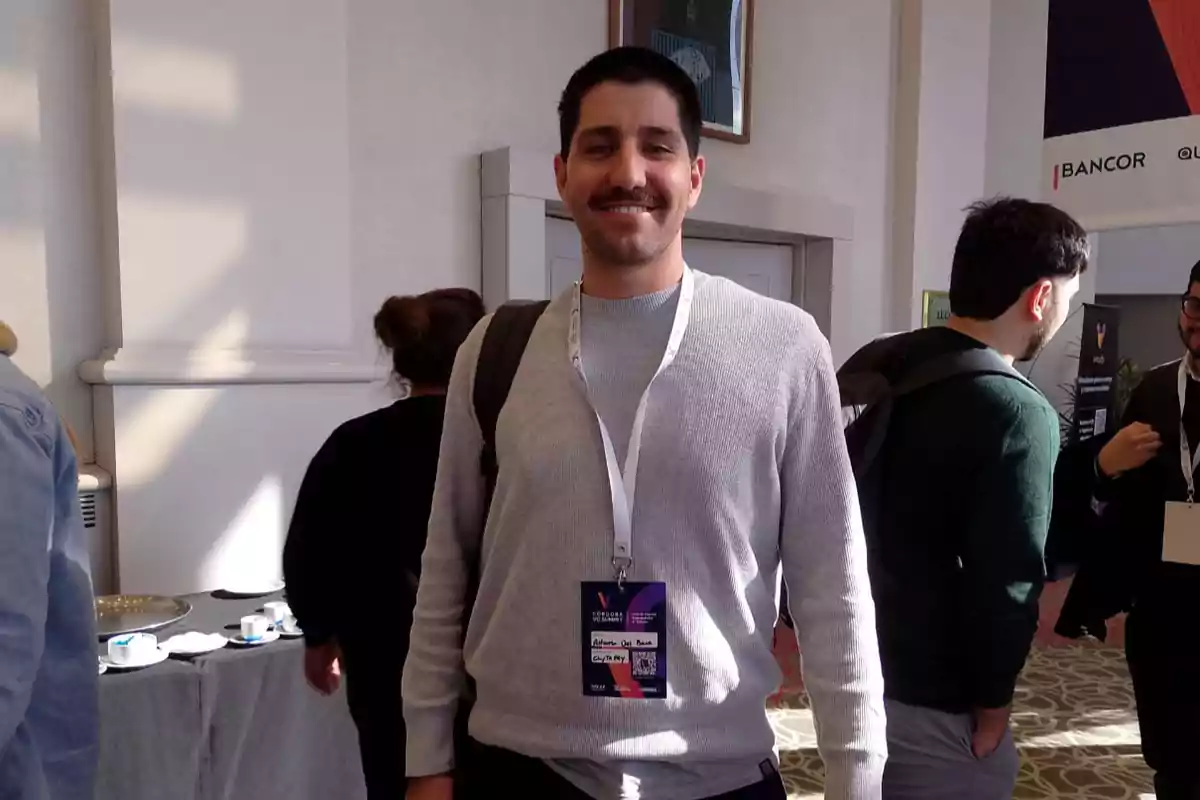 Young man with a mustache and gray sweater smiles while carrying a backpack and a badge at an event with other people in the background Young man with a mustache and gray sweater smiles while carrying a backpack and a badge at an event with other people in the background