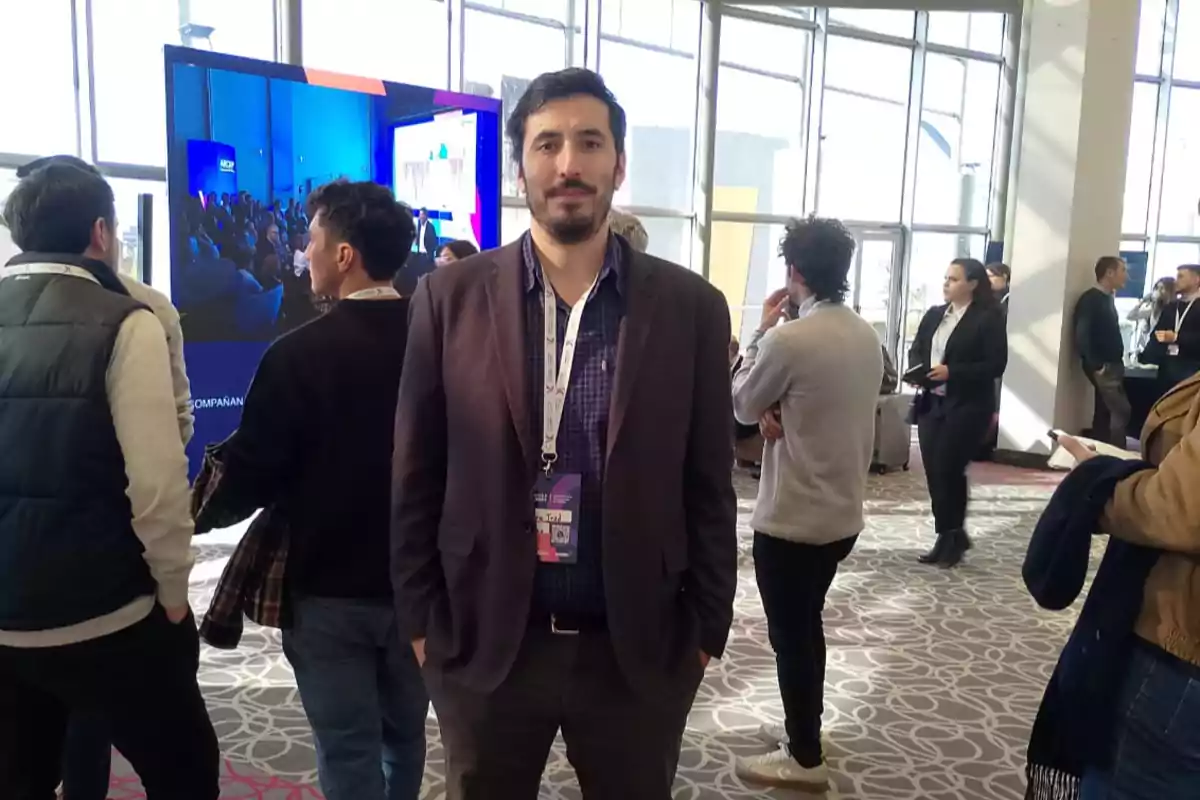 Man standing in the center of an event with several people around and a large screen in the background Man standing in the center of an event with several people around and a large screen in the background