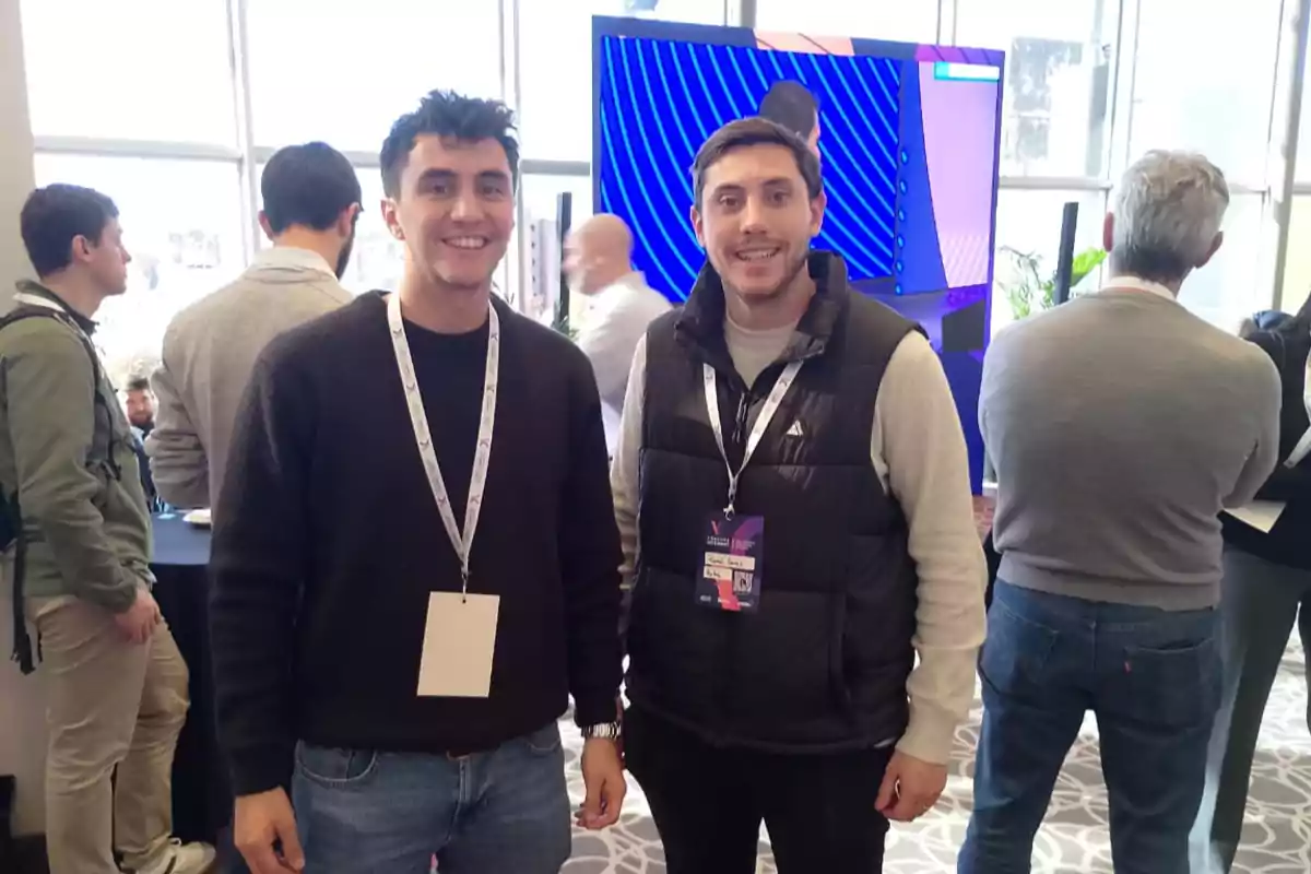 Two young men smile at the camera at an event with several people in the background and a large screen turned on. Two young men smile at the camera at an event with several people in the background and a large screen turned on.