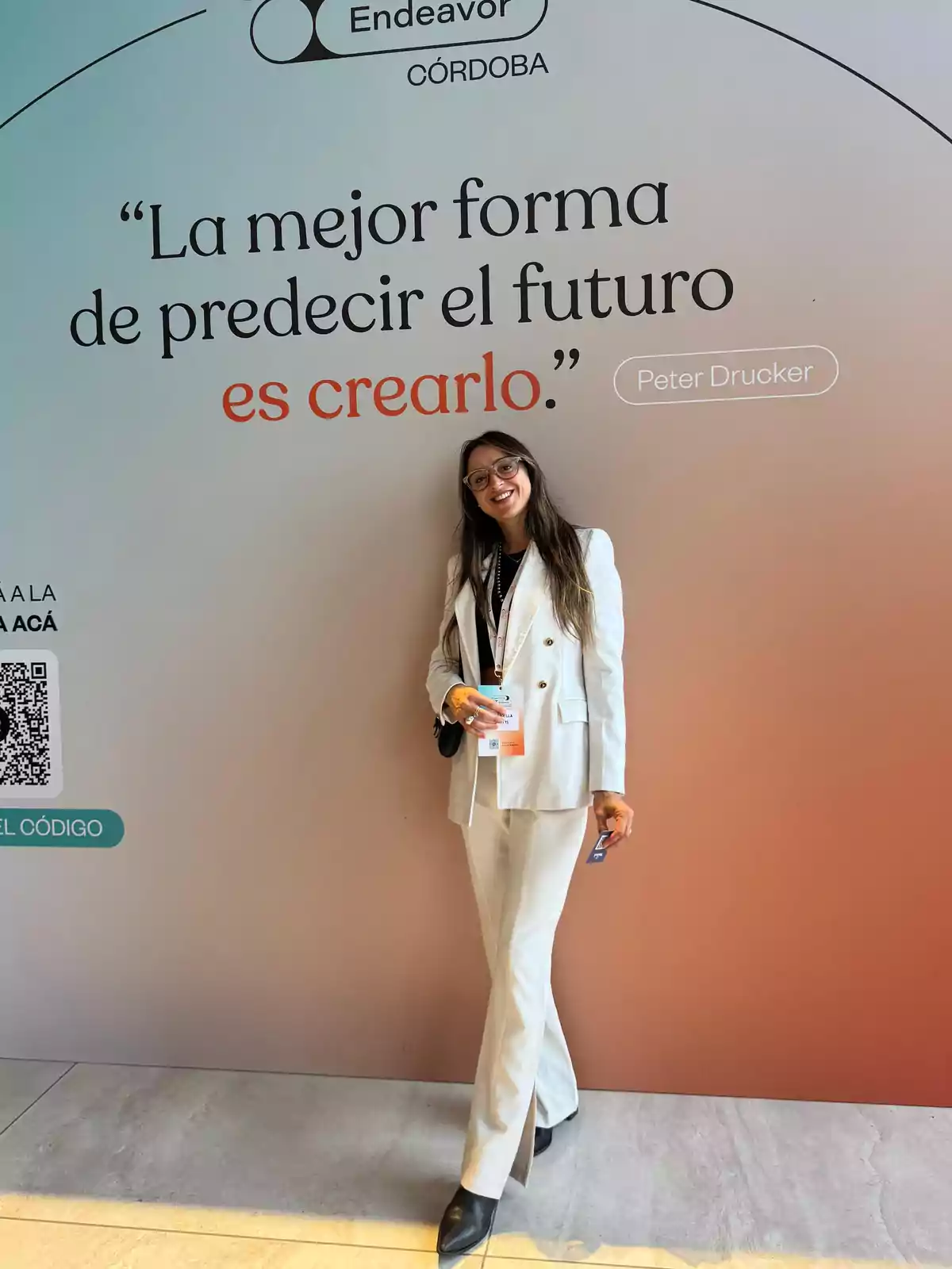 Mujer sonriente con gafas y traje blanco posando frente a un mural con una frase inspiradora sobre crear el futuro en un evento de Endeavor Córdoba Mujer sonriente con gafas y traje blanco posando frente a un mural con una frase inspiradora sobre crear el futuro en un evento de Endeavor Córdoba