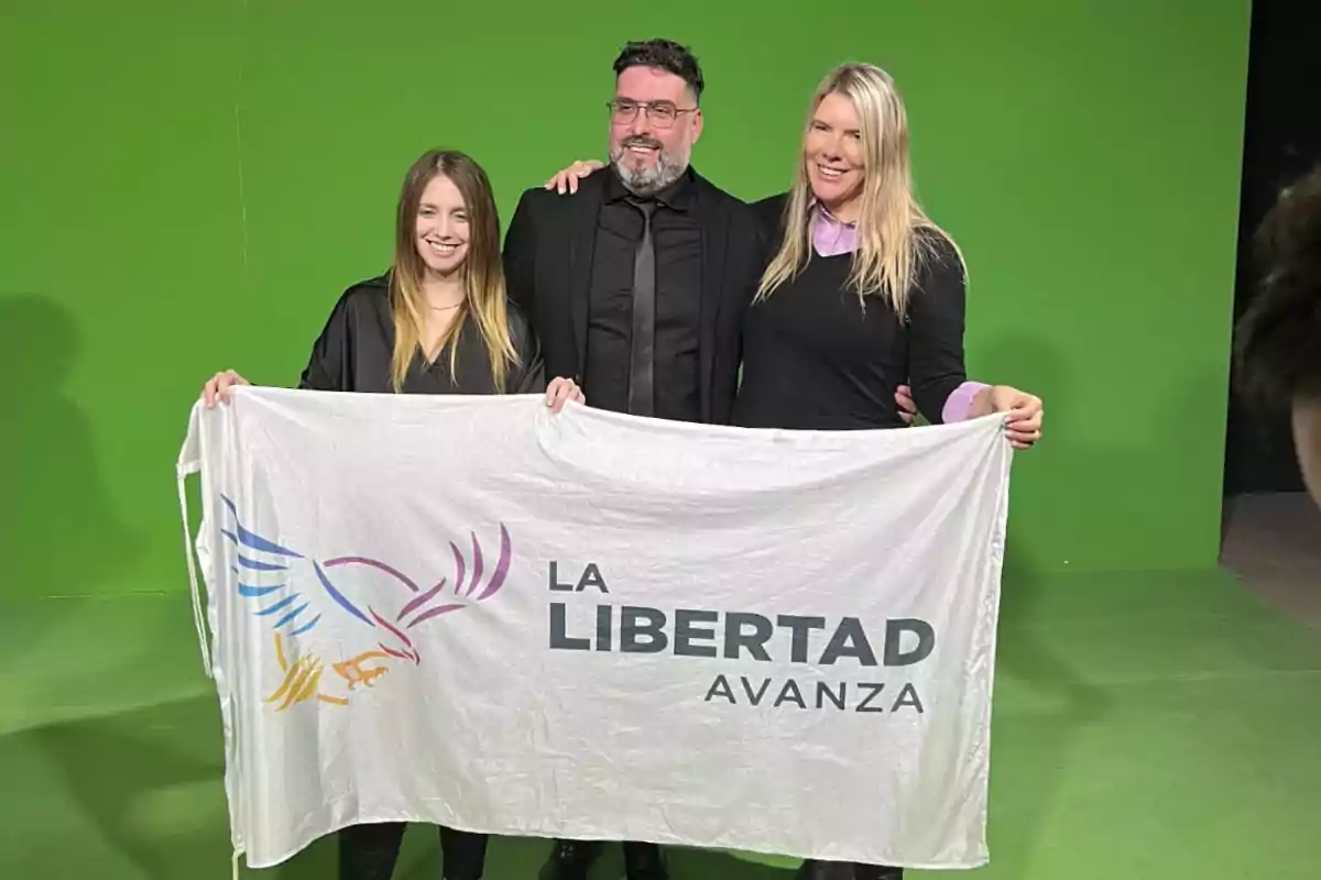 Tres personas sonríen frente a un fondo verde mientras sostienen una bandera blanca con el logo de un águila y el texto La Libertad Avanza.