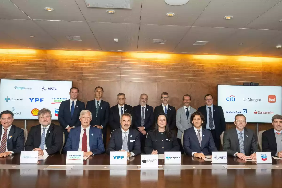 Grupo de ejecutivos y representantes de empresas energéticas y financieras posando en una sala de reuniones con pantallas que muestran logotipos de compañías reconocidas