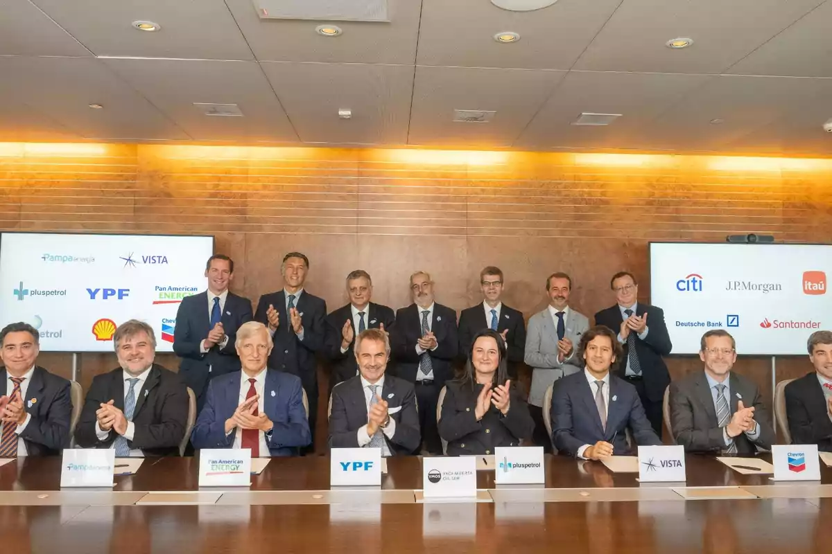 A group of people dressed in suits pose and applaud in a meeting room with two screens in the background displaying company and bank logos, while there are signs with the names of energy companies on the table. A group of people dressed in suits pose and applaud in a meeting room with two screens in the background displaying company and bank logos, while there are signs with the names of energy companies on the table.
