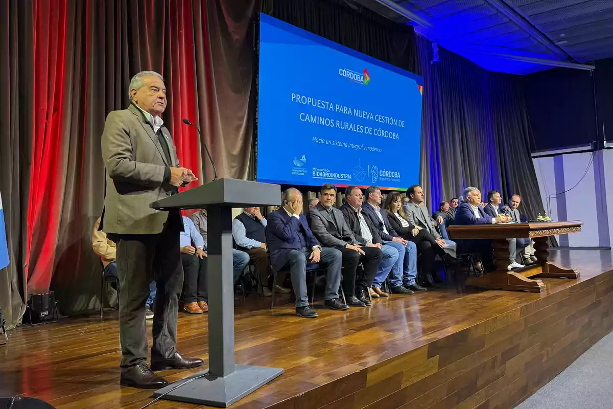 A man in a suit speaks at a podium in front of a group of people seated on a stage, with a blue screen in the background displaying a presentation about rural road management in Córdoba. A man in a suit speaks at a podium in front of a group of people seated on a stage, with a blue screen in the background displaying a presentation about rural road management in Córdoba.