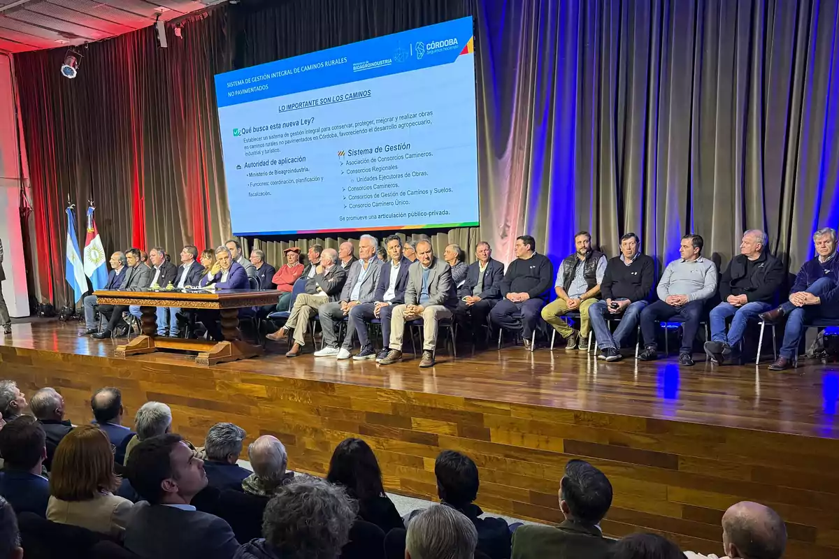 A large group of people seated on a stage during a formal event, with a big screen in the background displaying information about rural road management and Argentine flags to the side, while the audience watches from their seats.