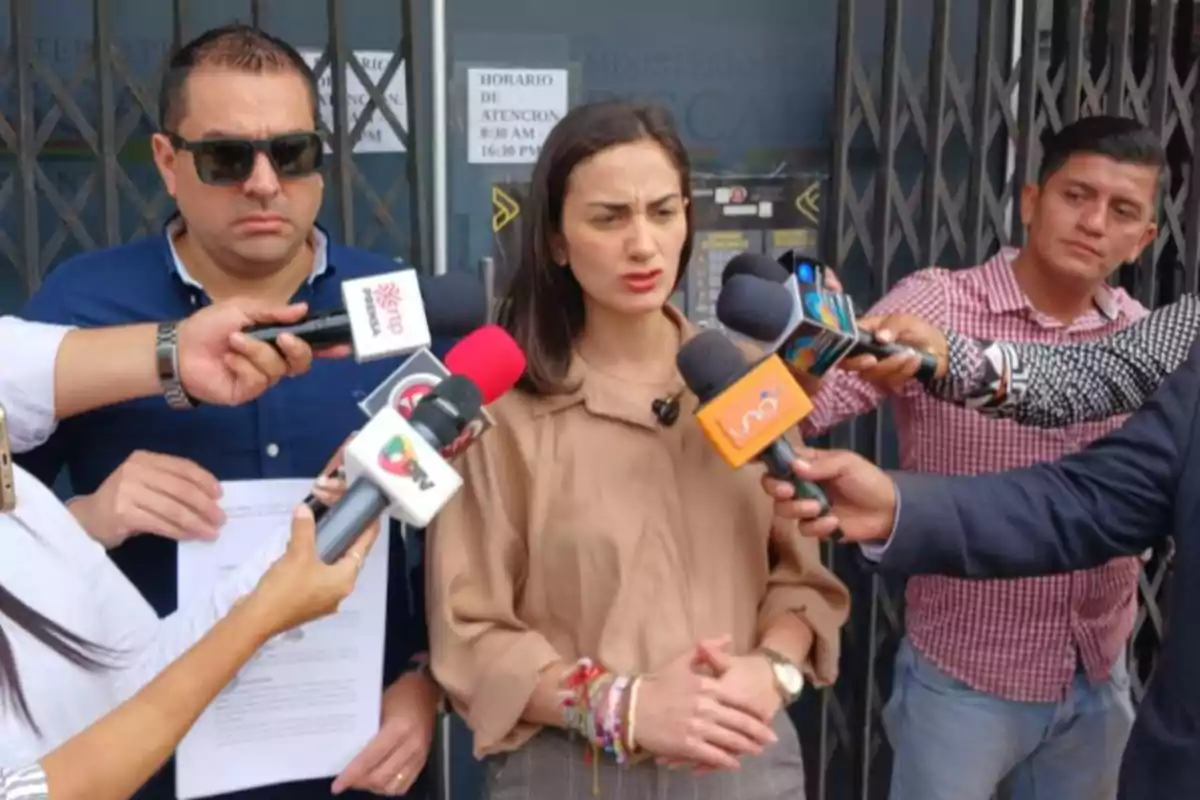 Una mujer da declaraciones a varios medios de comunicación mientras un hombre sostiene un documento a su lado y varias personas le acercan micrófonos. Una mujer da declaraciones a varios medios de comunicación mientras un hombre sostiene un documento a su lado y varias personas le acercan micrófonos.