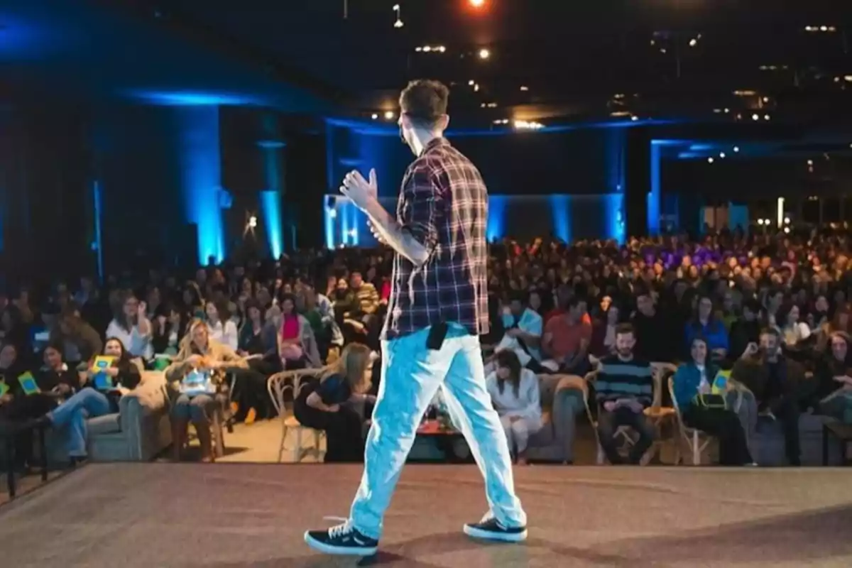 Man with his back to the audience giving a lecture on a stage in front of a large audience in an auditorium lit with blue lights