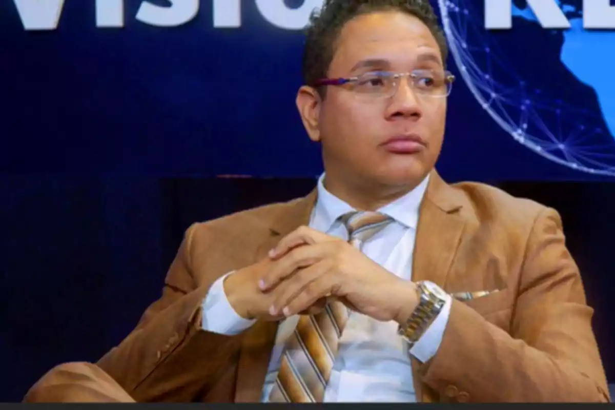 Hombre con gafas y traje marrón sentado con las manos entrelazadas frente a un fondo azul con letras blancas
