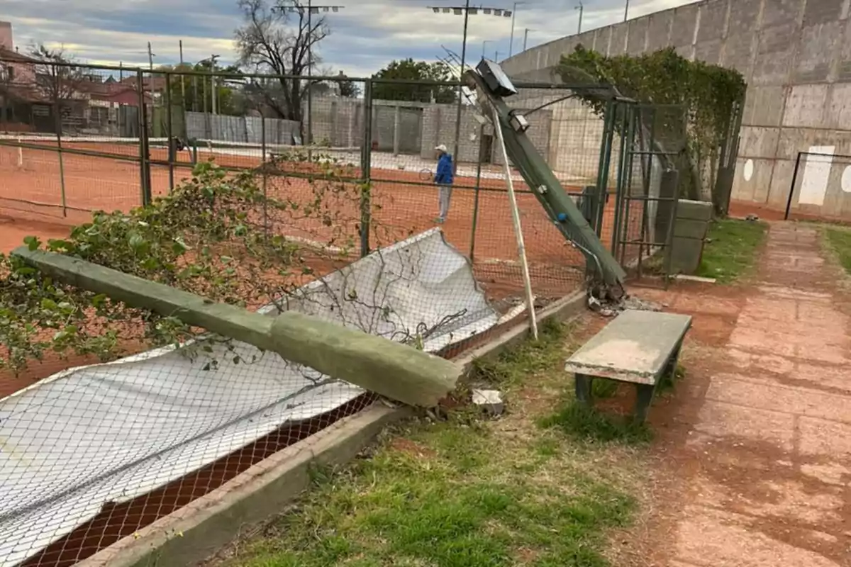 A fallen light pole on a fence in a tennis court with a person in the background