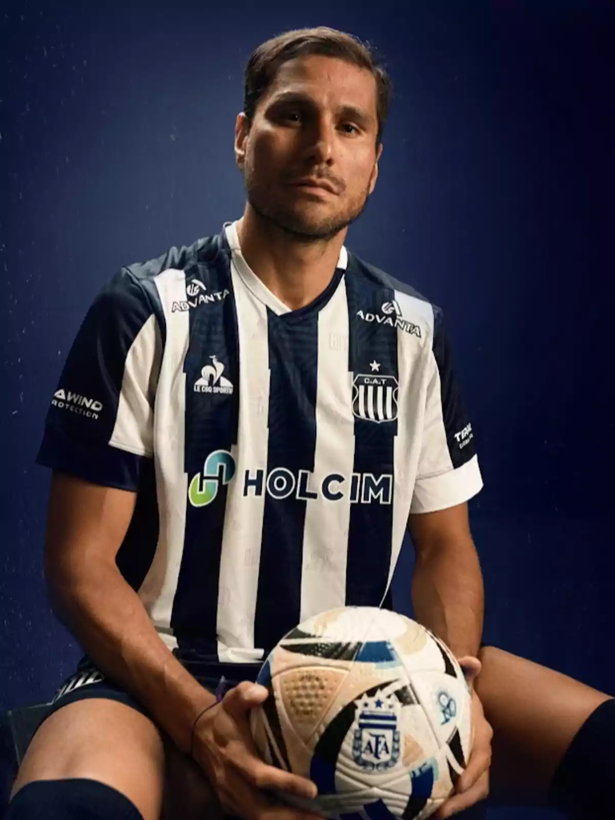 Futbolista con uniforme de Talleres de Córdoba sentado y sosteniendo un balón de fútbol frente a un fondo azul Futbolista con uniforme de Talleres de Córdoba sentado y sosteniendo un balón de fútbol frente a un fondo azul
