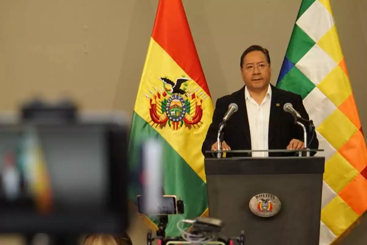 A man in a dark suit and white jersey gives a speech at a podium in front of the flags of Bolivia and the Wiphala while being recorded by cameras.