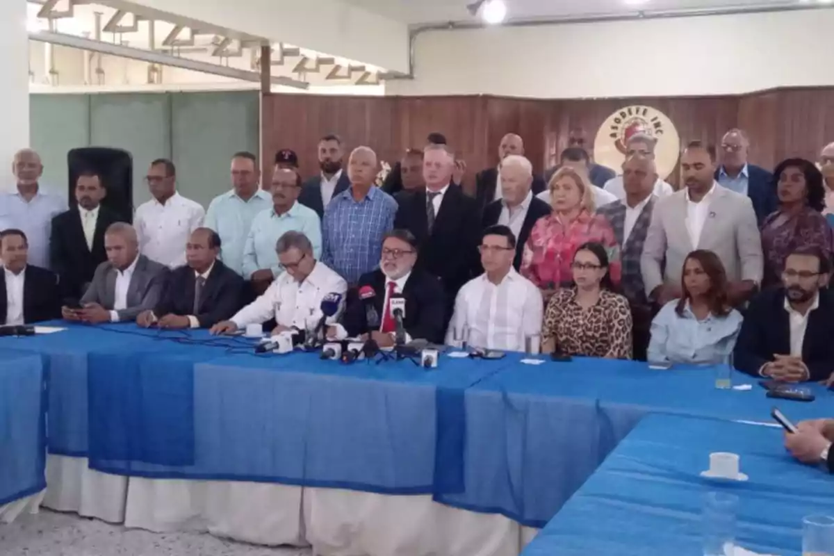 Un grupo numeroso de personas, algunas sentadas y otras de pie, participa en una rueda de prensa en un salón con mesas cubiertas con manteles azules y varios micrófonos frente a los oradores principales.