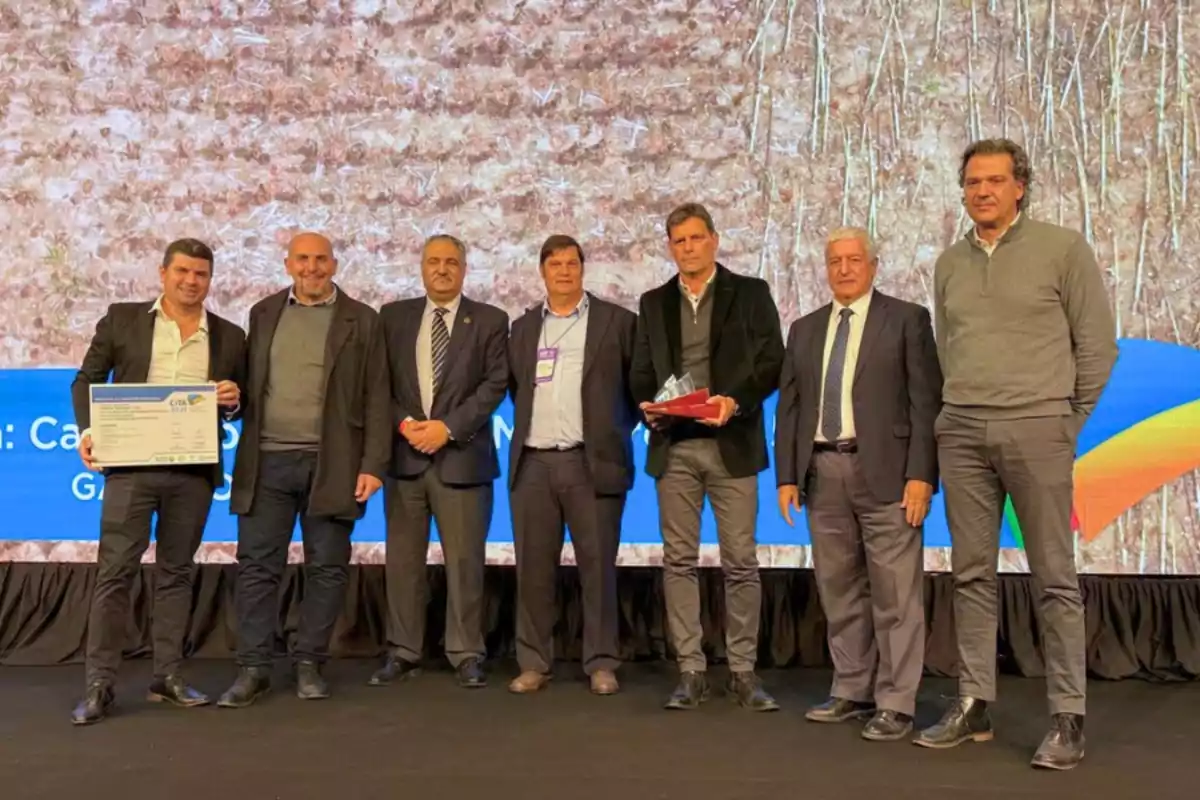 Siete hombres posan en un escenario durante un evento, dos de ellos sostienen un diploma y un trofeo, todos visten ropa formal y de negocios.