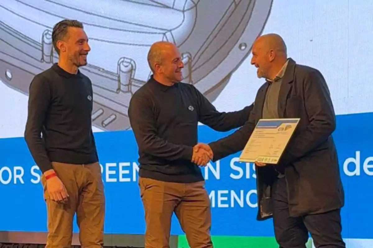 Tres hombres sonríen mientras uno de ellos entrega un reconocimiento a otro en un escenario durante un evento Tres hombres sonríen mientras uno de ellos entrega un reconocimiento a otro en un escenario durante un evento