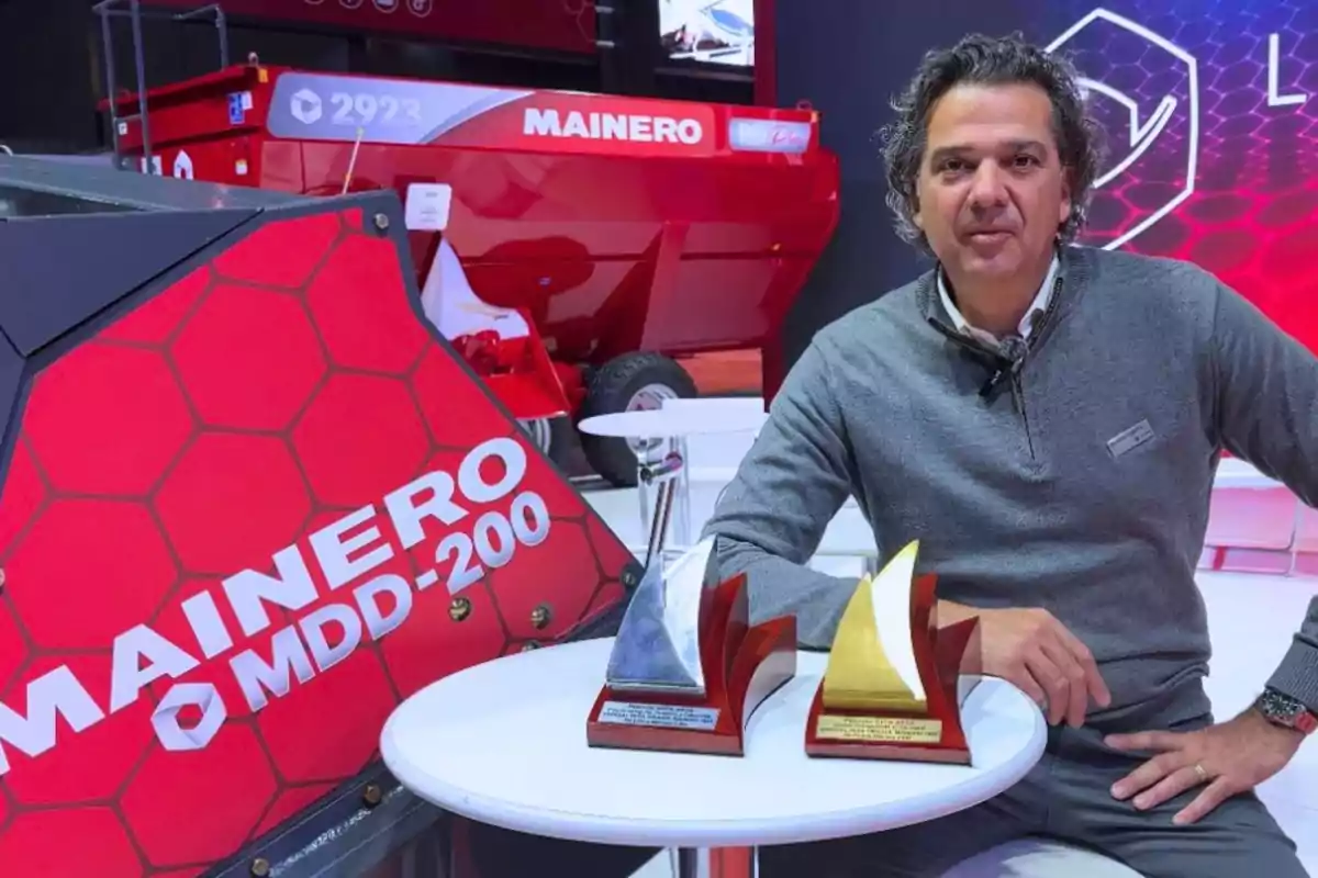 Hombre sentado en una mesa blanca junto a dos trofeos, con maquinaria agrícola roja de la marca Mainero al fondo Hombre sentado en una mesa blanca junto a dos trofeos, con maquinaria agrícola roja de la marca Mainero al fondo