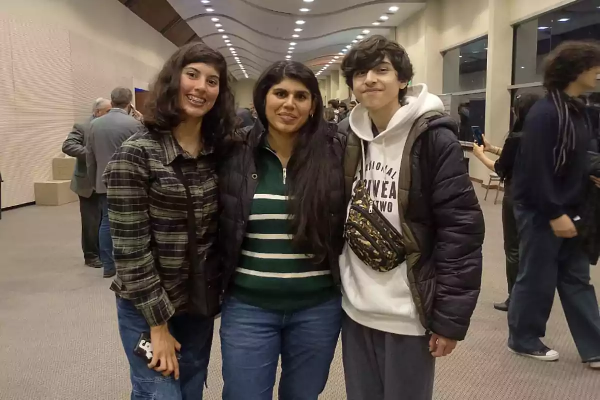 Tres personas jóvenes posan sonrientes en el centro de un salón amplio y bien iluminado, con otras personas conversando al fondo.
