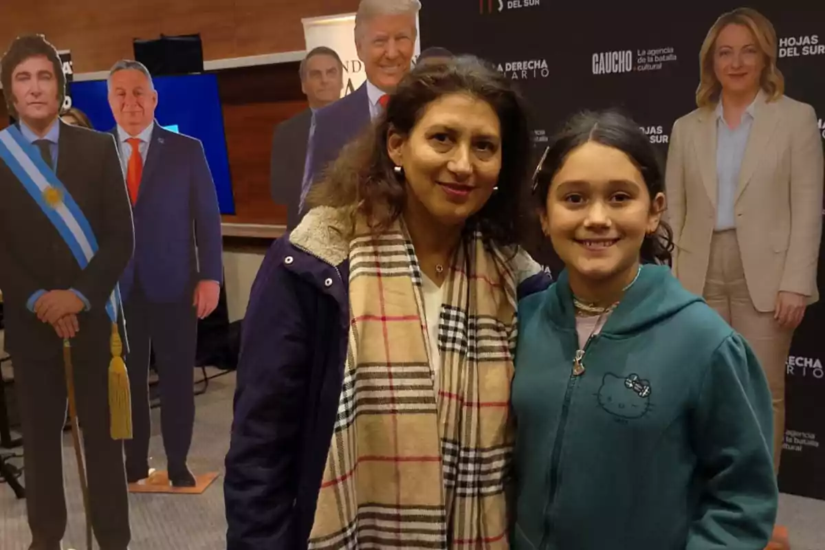 Dos personas sonríen frente a la cámara en un evento, detrás de ellas hay figuras de cartón de políticos reconocidos y un fondo con logotipos.