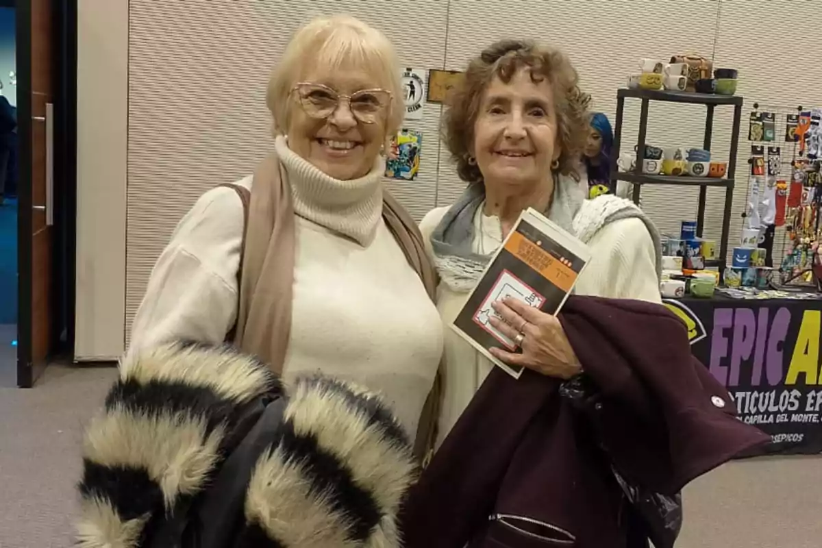 Dos mujeres mayores sonríen a la cámara en un evento bajo techo una de ellas sostiene un libro y ambas llevan ropa de abrigo al fondo se observa un puesto con tazas y otros artículos.