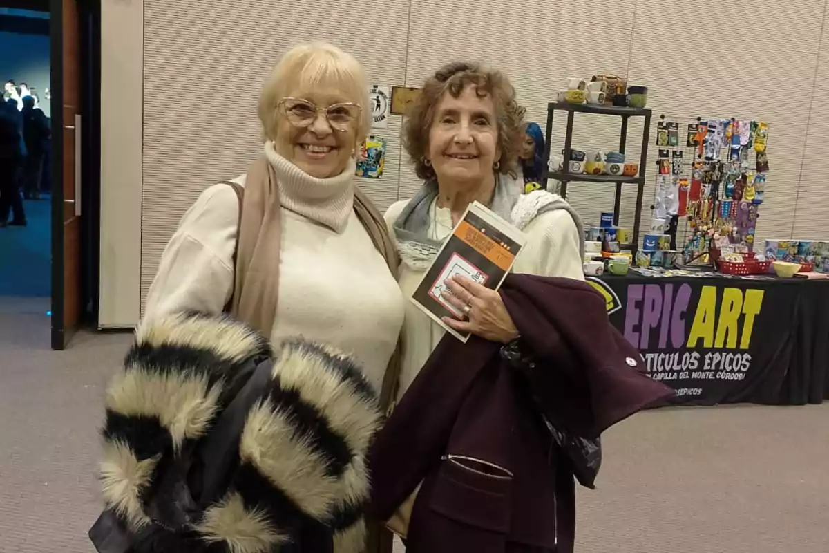 Dos mujeres mayores sonríen a la cámara en un evento, una sostiene un libro y la otra un abrigo de peluche, al fondo hay un puesto de artículos coloridos llamado Epic Art.