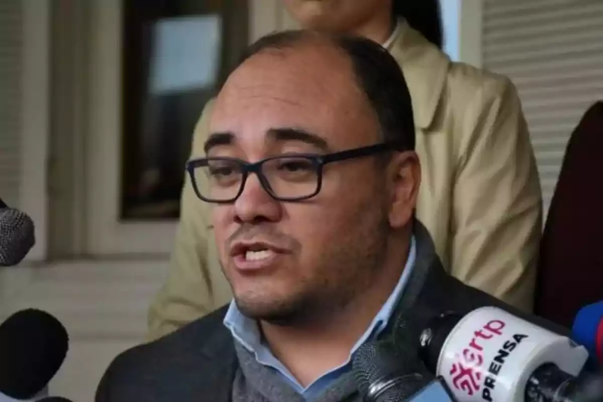 Hombre con gafas hablando frente a varios micrófonos en una conferencia de prensa Hombre con gafas hablando frente a varios micrófonos en una conferencia de prensa