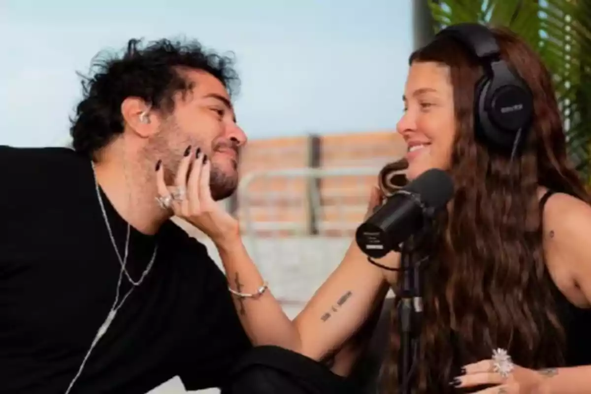 Two people smile and look at each other affectionately while recording a podcast; one of them is wearing large headphones and holding the other’s face with their hand.