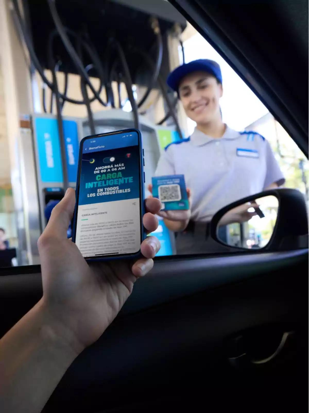 Persona dentro de un auto sostiene un celular con una promoción de carga inteligente de combustible mientras una empleada de estación de servicio sonríe y muestra un código QR junto a un surtidor Persona dentro de un auto sostiene un celular con una promoción de carga inteligente de combustible mientras una empleada de estación de servicio sonríe y muestra un código QR junto a un surtidor