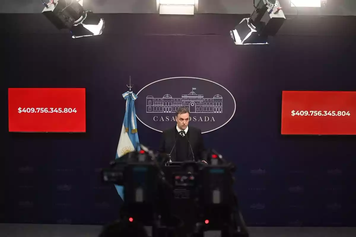 A man in a suit gives a press conference at the Casa Rosada with two red screens on either side displaying the number 409,756,345,804 and an Argentine flag next to him.