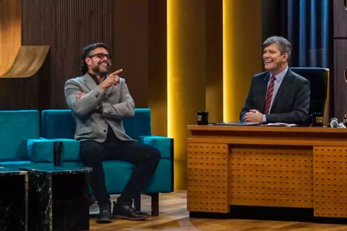 Dos hombres sonríen y conversan en un set de programa de entrevistas uno sentado en un sillón azul y el otro detrás de un escritorio