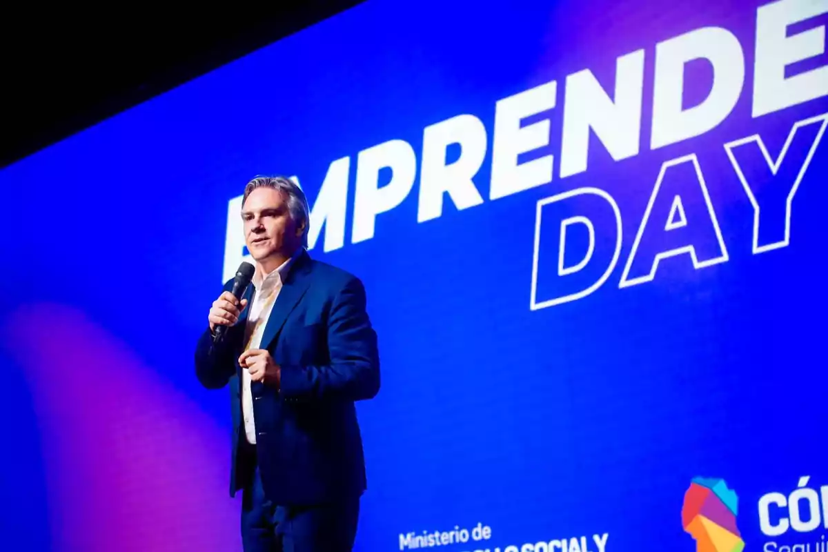 Hombre de traje azul hablando con micrófono en mano frente a una pantalla azul que dice EMPRENDE DAY Hombre de traje azul hablando con micrófono en mano frente a una pantalla azul que dice EMPRENDE DAY