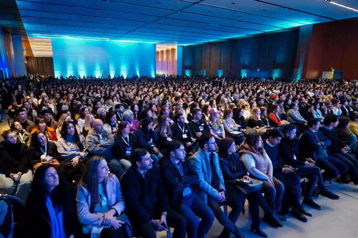 Auditorio lleno de personas sentadas prestando atención a un evento en un salón iluminado con luces azules