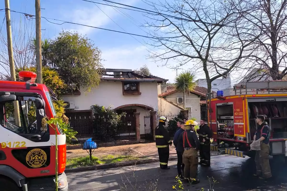 Bomberos y camiones de bomberos frente a una casa que muestra daños por incendio en el techo mientras conversan en la calle