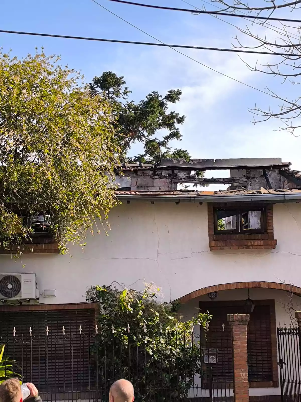 Two-story house with visible damage to the roof and walls, surrounded by vegetation and with people watching from the street. Two-story house with visible damage to the roof and walls, surrounded by vegetation and with people watching from the street.