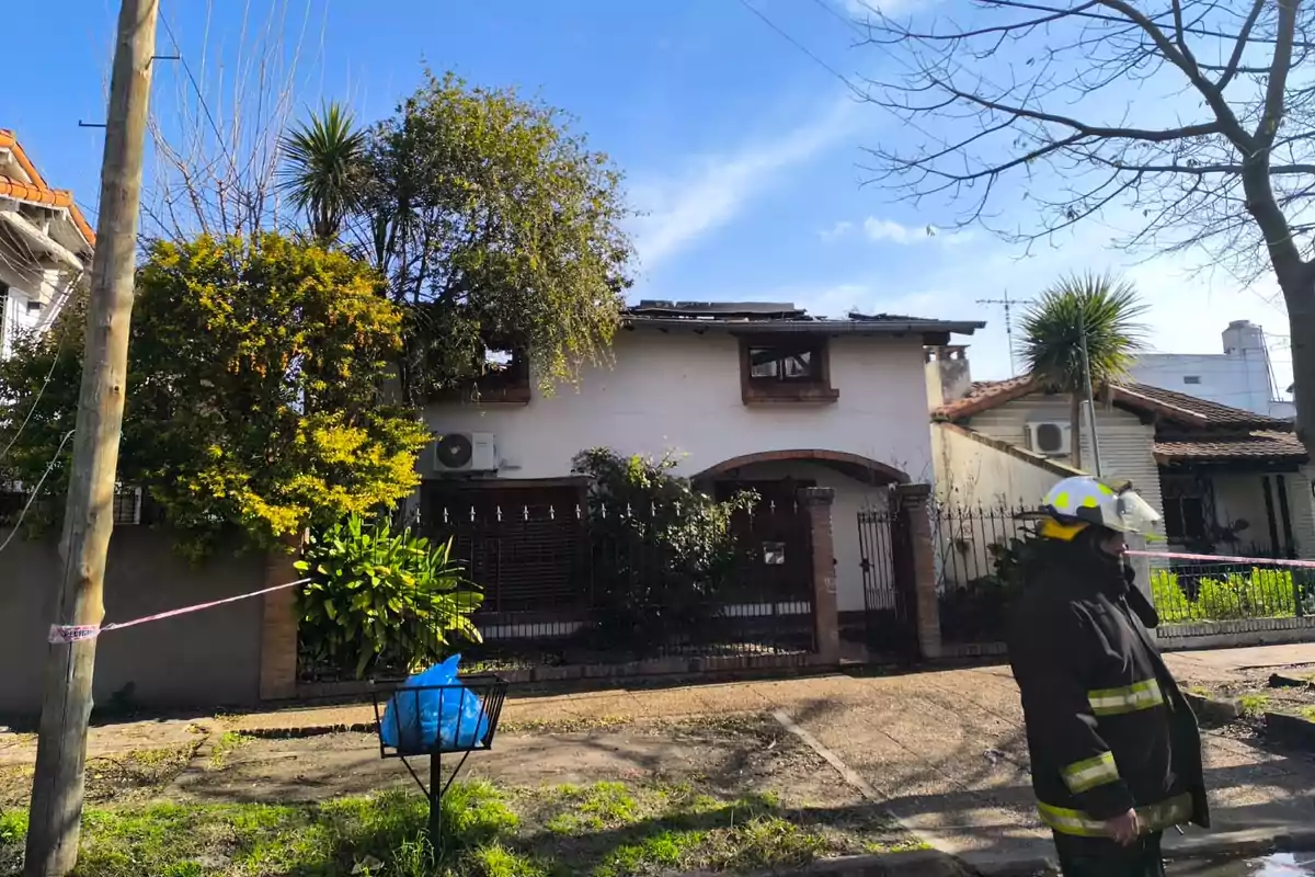 Two-story house with visible roof damage, surrounded by trees and plants, with a firefighter standing on the sidewalk in front of the home on a sunny day. Two-story house with visible roof damage, surrounded by trees and plants, with a firefighter standing on the sidewalk in front of the home on a sunny day.