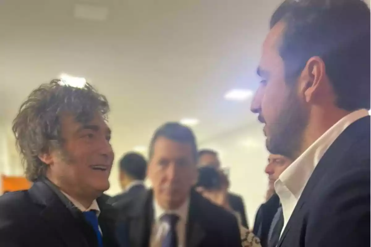 Dos hombres conversan sonrientes en un ambiente interior con varias personas al fondo Dos hombres conversan sonrientes en un ambiente interior con varias personas al fondo