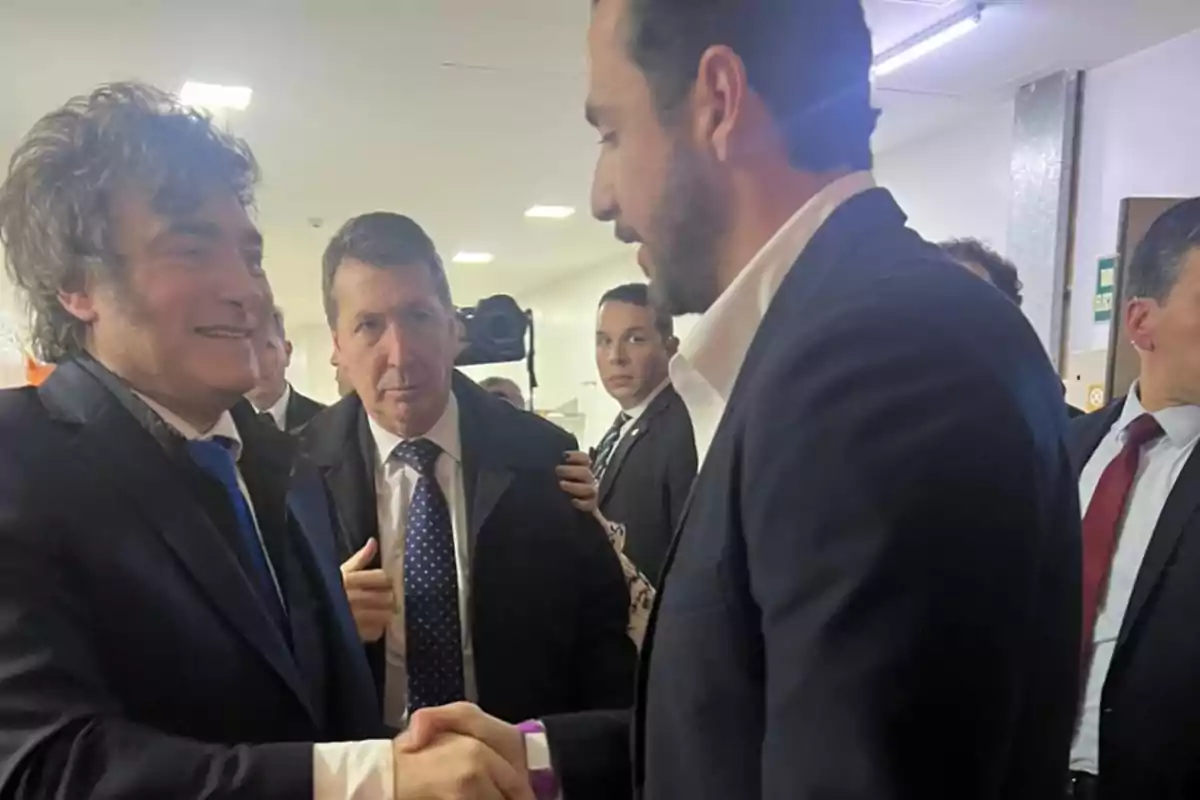 Dos hombres vestidos de traje se dan la mano mientras otras personas los observan en un pasillo iluminado.