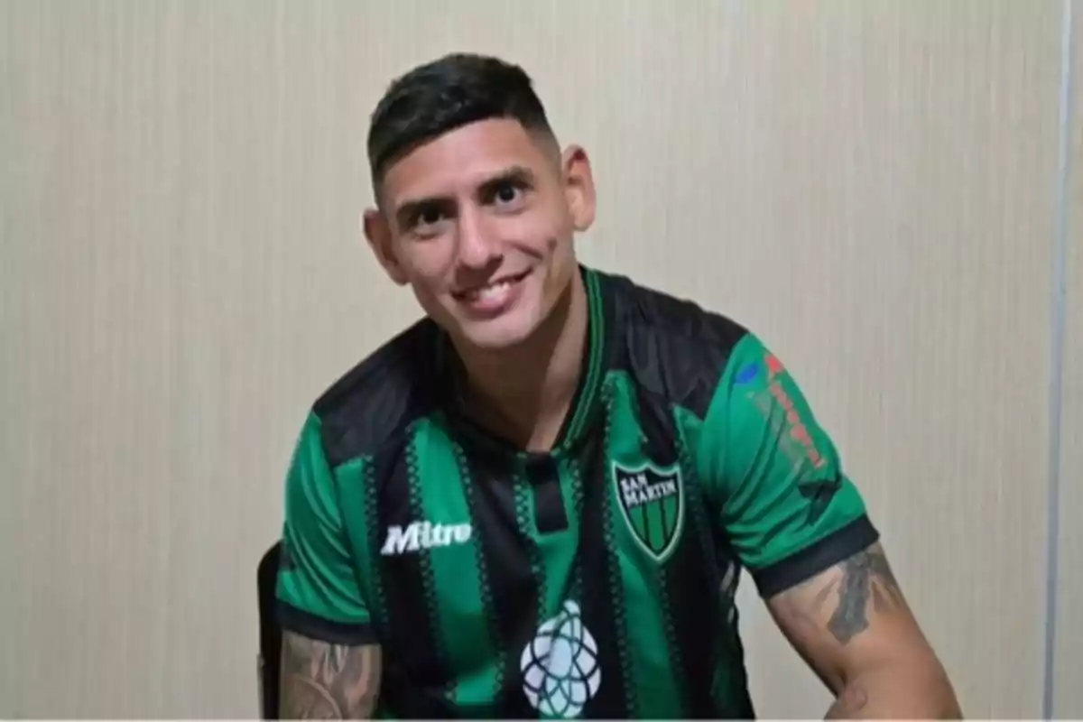 Hombre con camiseta de fútbol verde y negra con el escudo de San Martín sentado y sonriendo frente a una pared clara