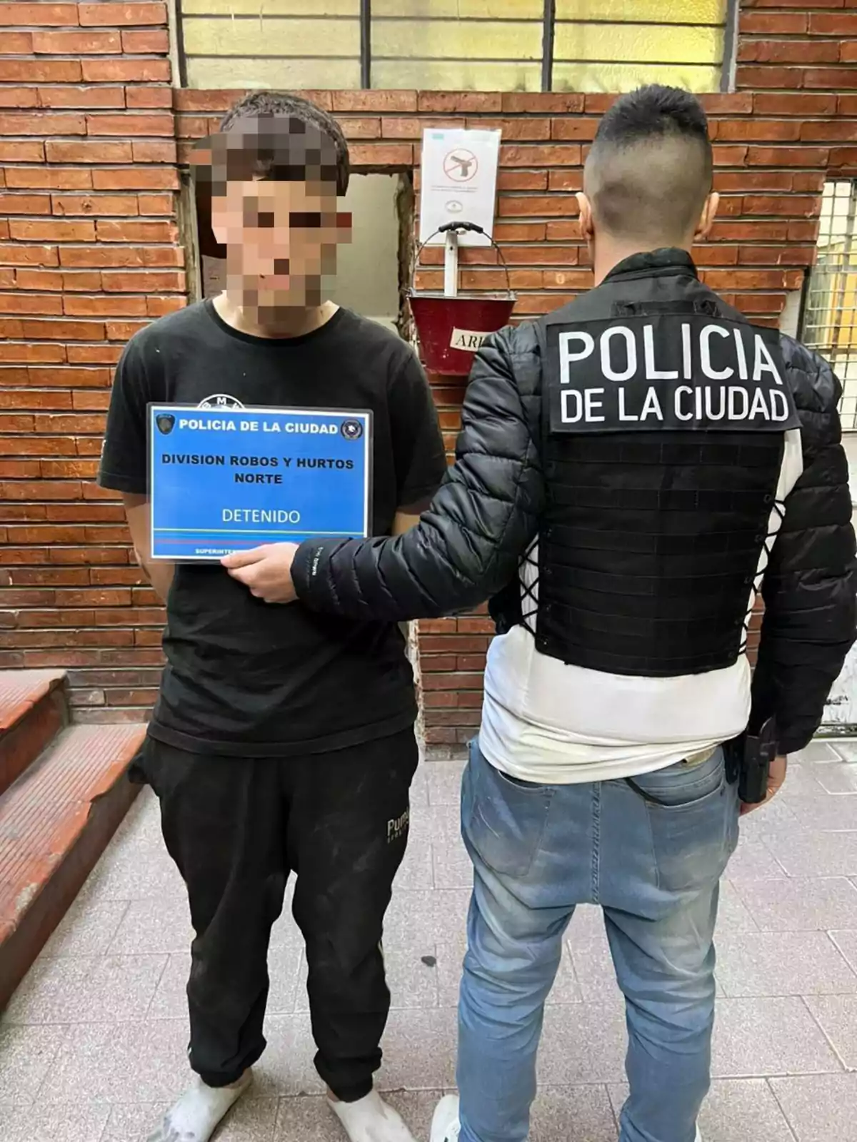 Un joven con el rostro pixelado sostiene un cartel de detenido junto a un policía de la ciudad que lleva una chaqueta identificatoria, ambos están de pie frente a una pared de ladrillos. Un joven con el rostro pixelado sostiene un cartel de detenido junto a un policía de la ciudad que lleva una chaqueta identificatoria, ambos están de pie frente a una pared de ladrillos.