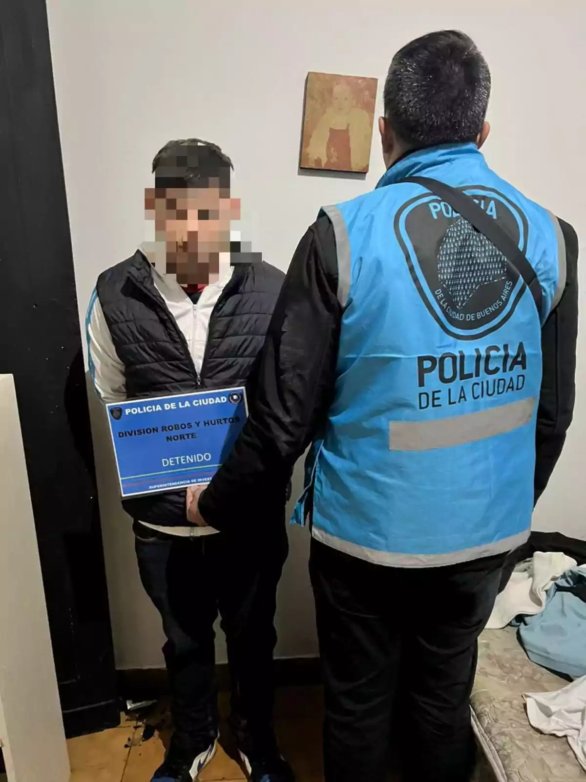Un hombre con el rostro pixelado sostiene un cartel de detenido mientras un oficial de policía con chaleco celeste le da la espalda en una habitación sencilla Un hombre con el rostro pixelado sostiene un cartel de detenido mientras un oficial de policía con chaleco celeste le da la espalda en una habitación sencilla