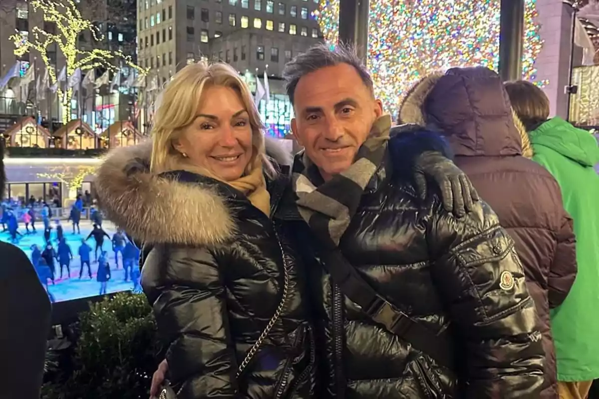 Pareja sonriente con abrigos de invierno posando frente a una pista de patinaje y un árbol de Navidad iluminado en una ciudad