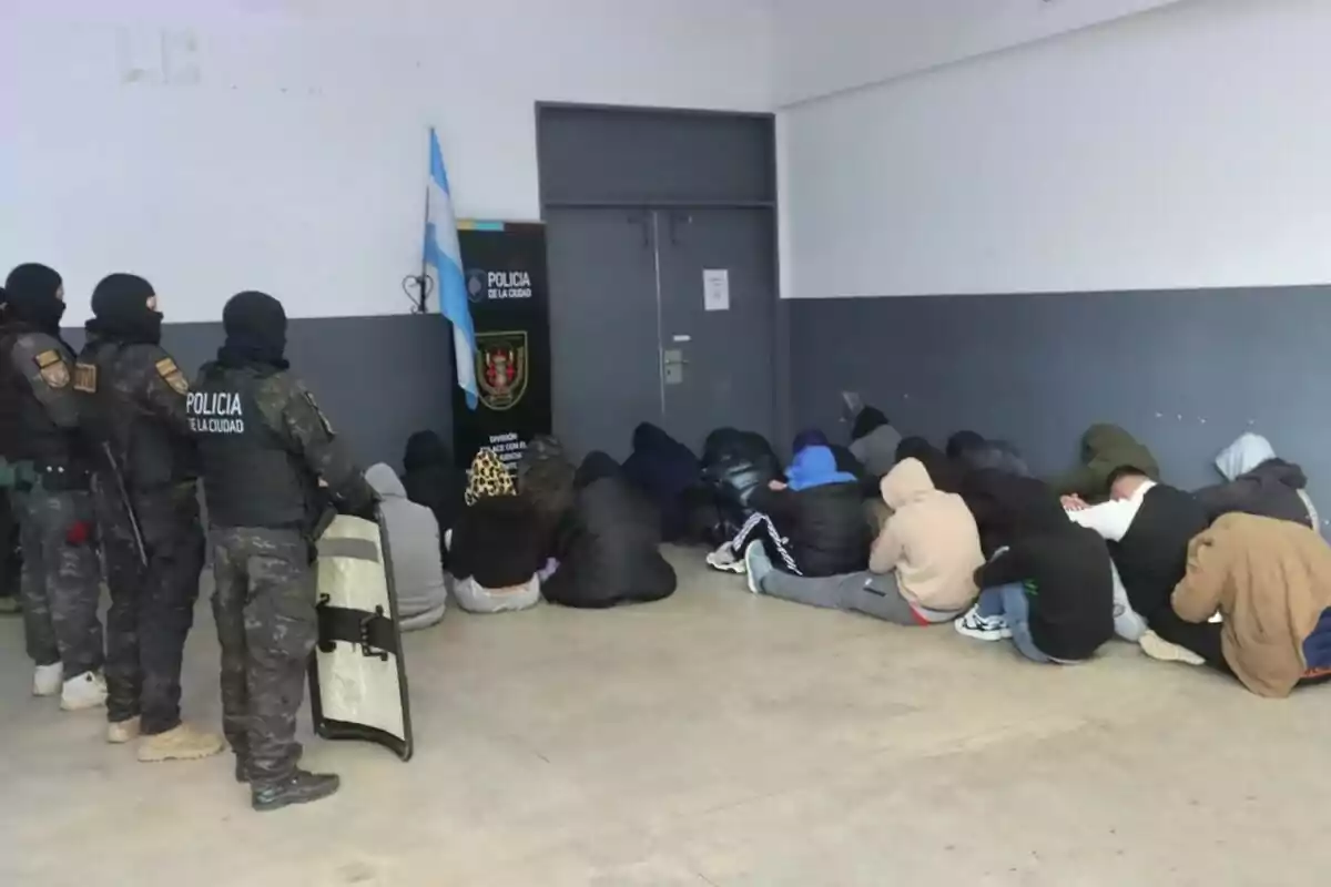 Several heavily armed police officers guard a group of people sitting on the floor with their heads down in a closed room. Several heavily armed police officers guard a group of people sitting on the floor with their heads down in a closed room.
