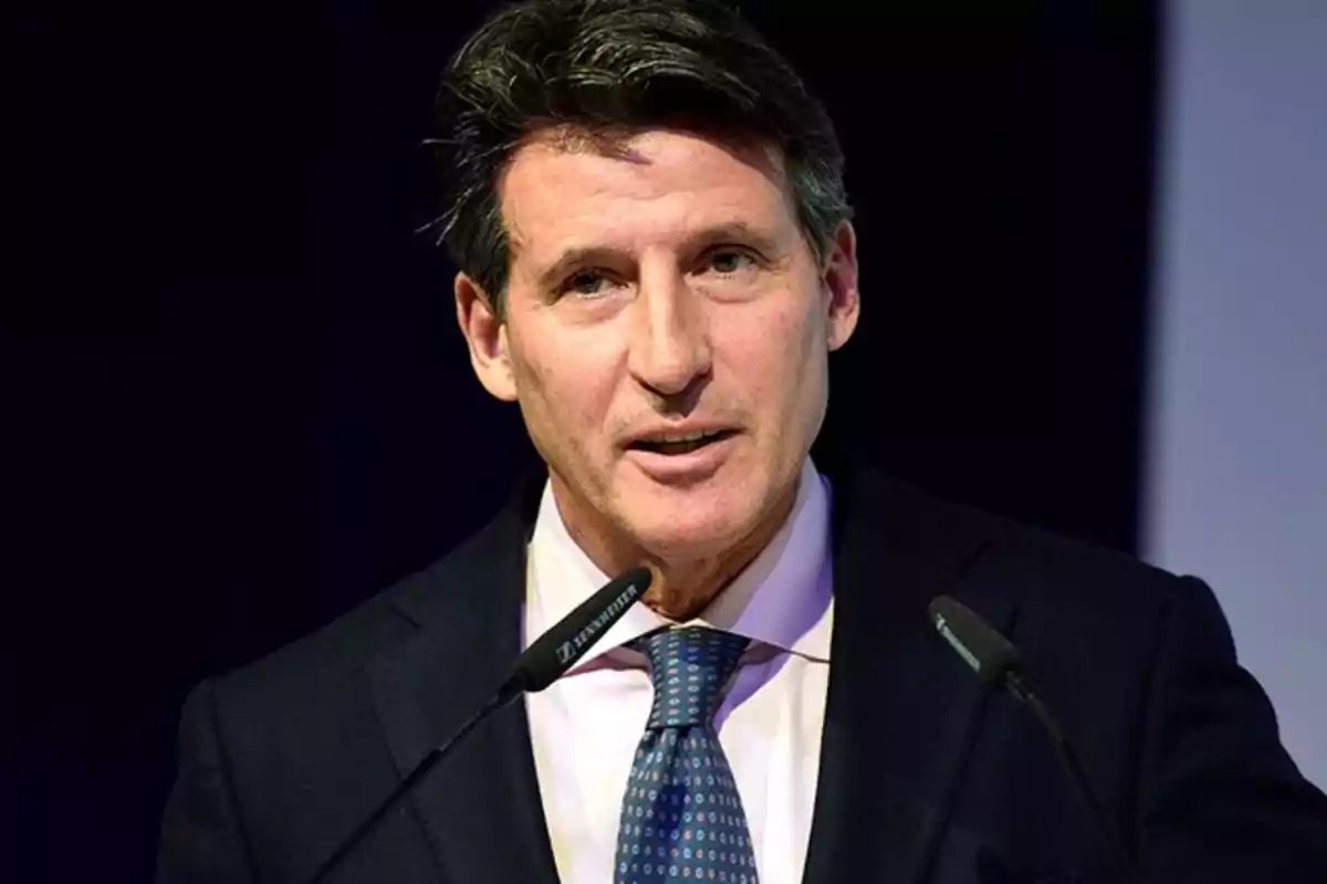 Hombre de cabello oscuro con traje y corbata hablando frente a dos micrófonos Hombre de cabello oscuro con traje y corbata hablando frente a dos micrófonos