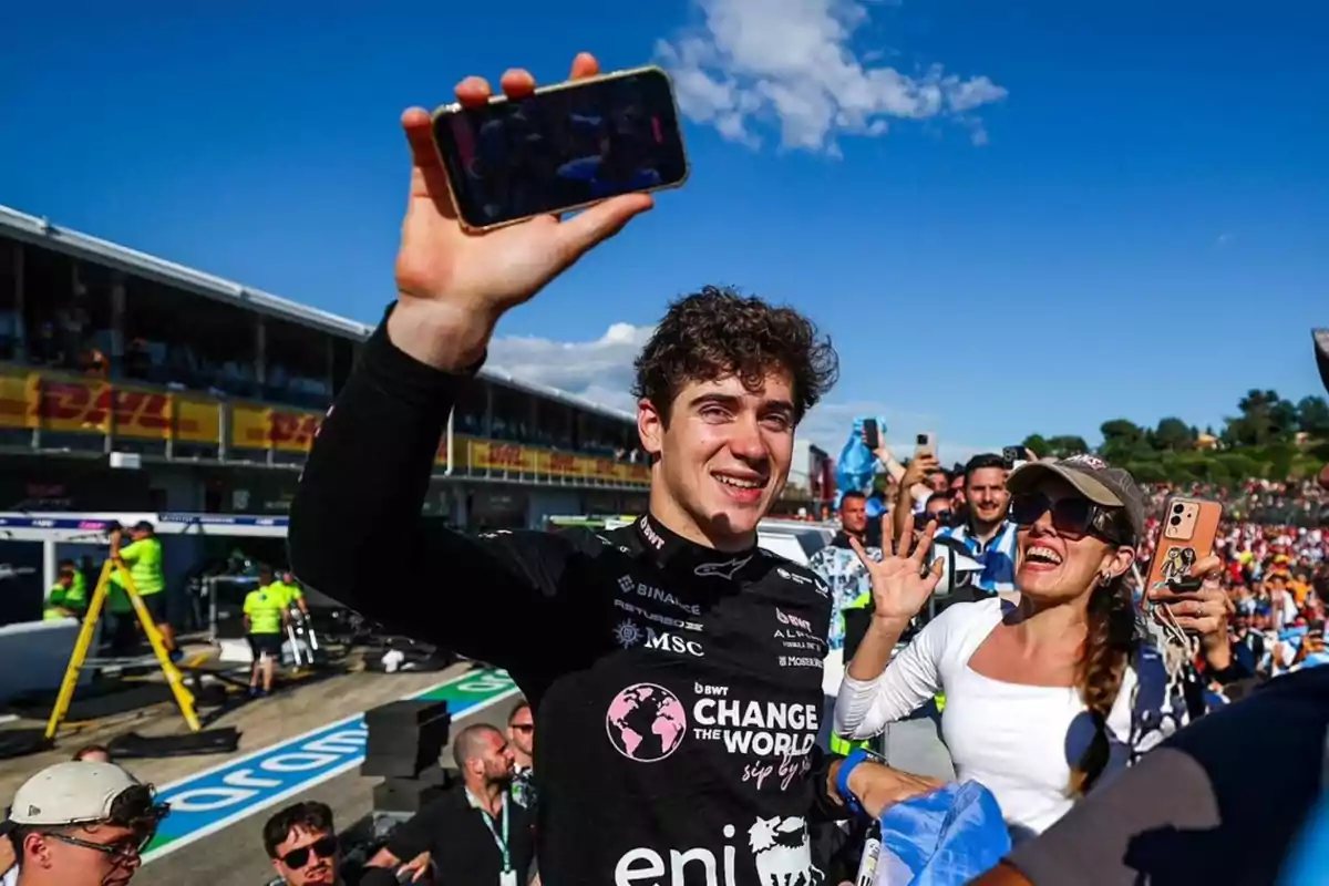Piloto de automovilismo sonriente celebrando con aficionados en un circuito mientras sostiene un teléfono móvil para tomarse una selfie Piloto de automovilismo sonriente celebrando con aficionados en un circuito mientras sostiene un teléfono móvil para tomarse una selfie