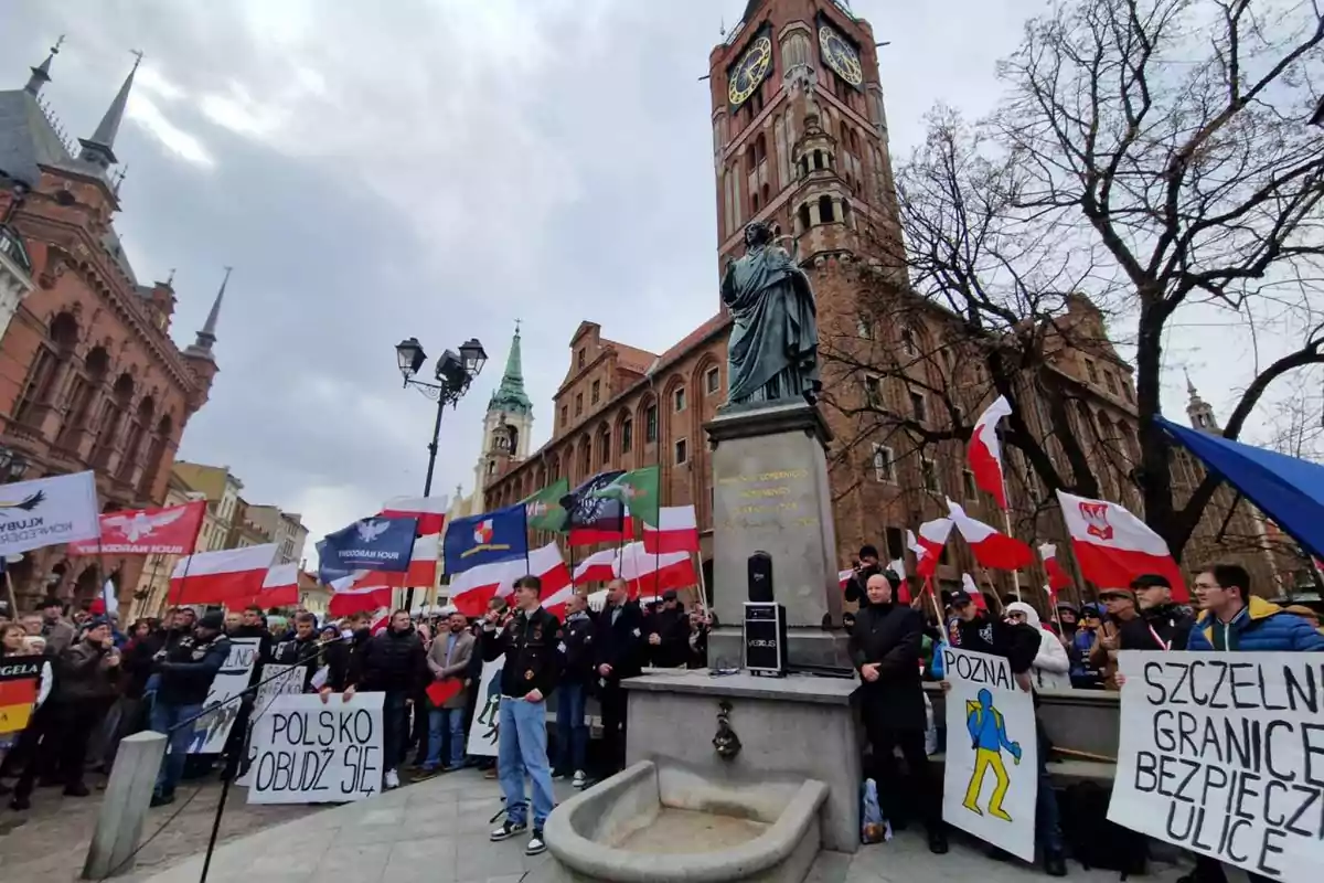 Demonstration in a square with many people holding Polish flags and banners, next to a statue and a historic brick building with a large clock. Demonstration in a square with many people holding Polish flags and banners, next to a statue and a historic brick building with a large clock.