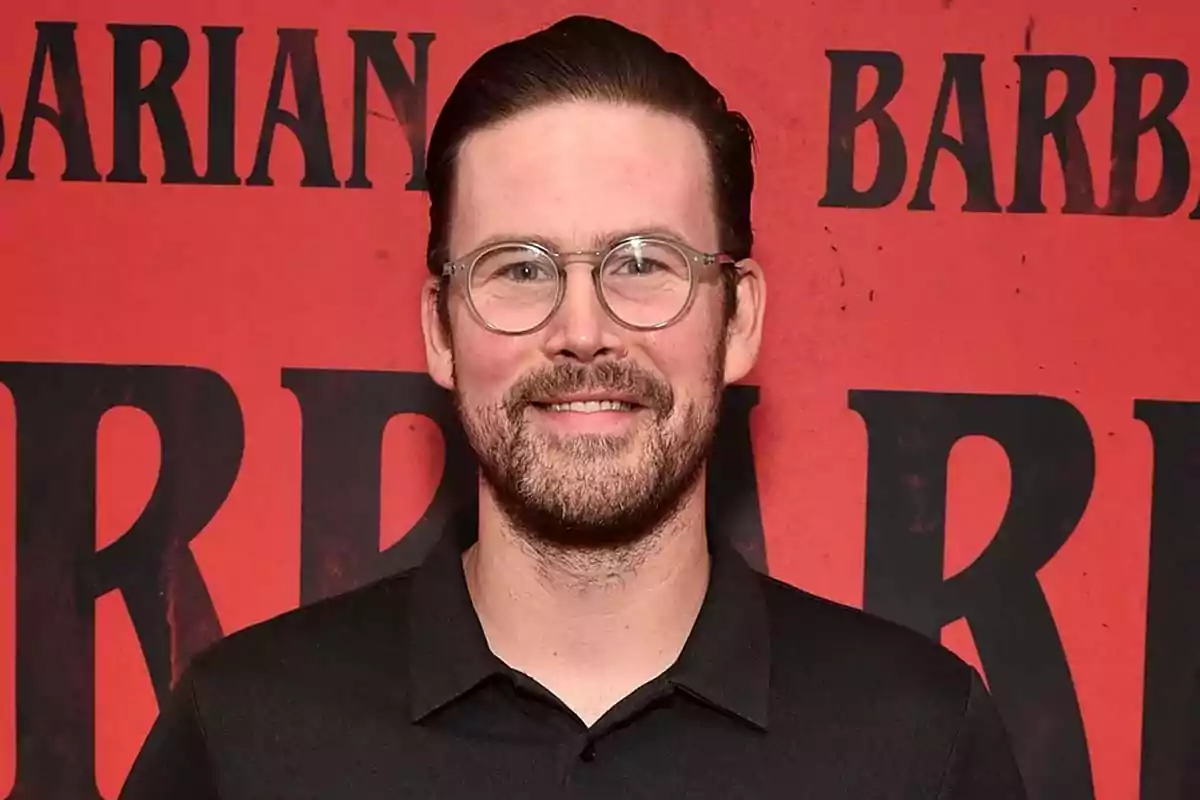 Hombre con gafas y barba sonríe frente a un fondo rojo con letras negras Hombre con gafas y barba sonríe frente a un fondo rojo con letras negras