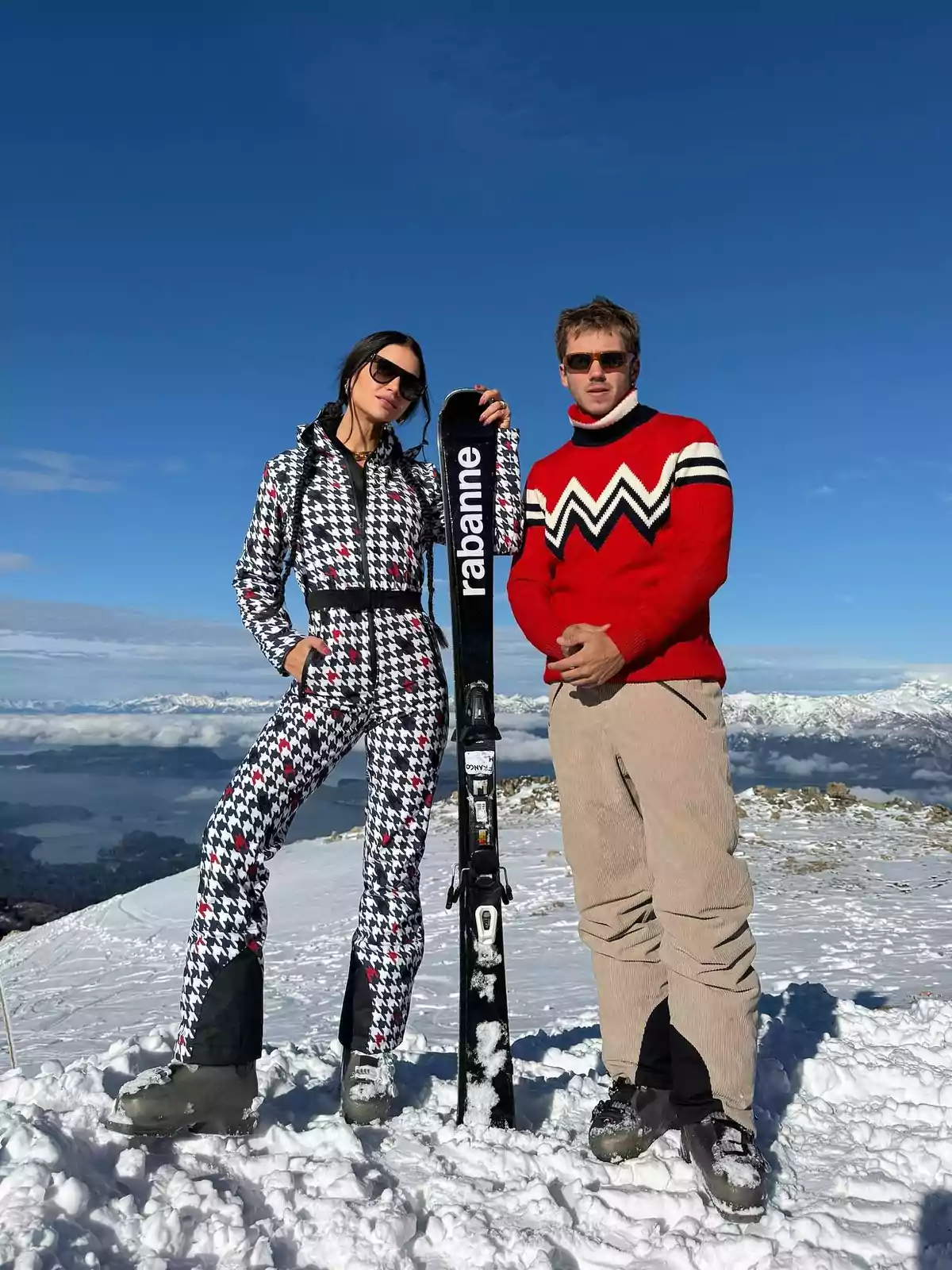 Dos personas posan en la nieve con ropa de esquí y gafas de sol, una de ellas sostiene un esquí negro con la palabra rabanne, el cielo está despejado y se ven montañas al fondo Dos personas posan en la nieve con ropa de esquí y gafas de sol, una de ellas sostiene un esquí negro con la palabra rabanne, el cielo está despejado y se ven montañas al fondo