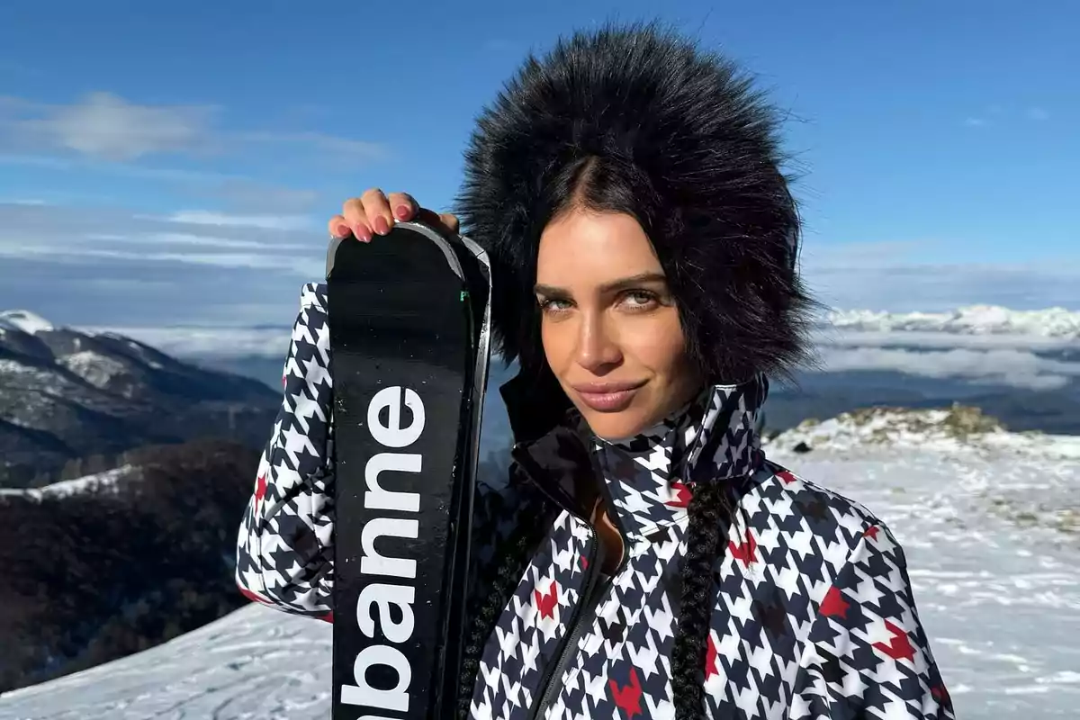 Mujer con abrigo de invierno y gorro de peluche negro sosteniendo un esquí en una montaña nevada con cielo azul de fondo
