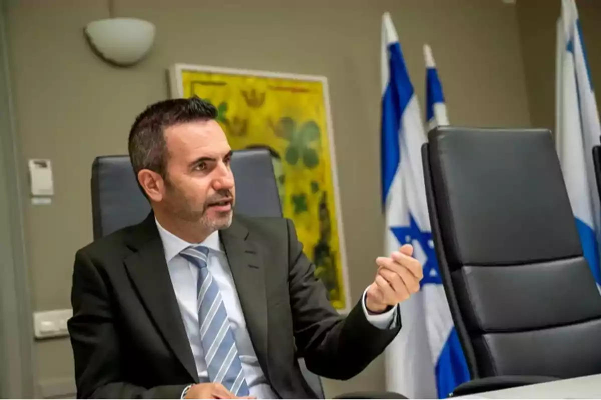 Hombre de traje sentado en una sala de reuniones con banderas de Israel al fondo