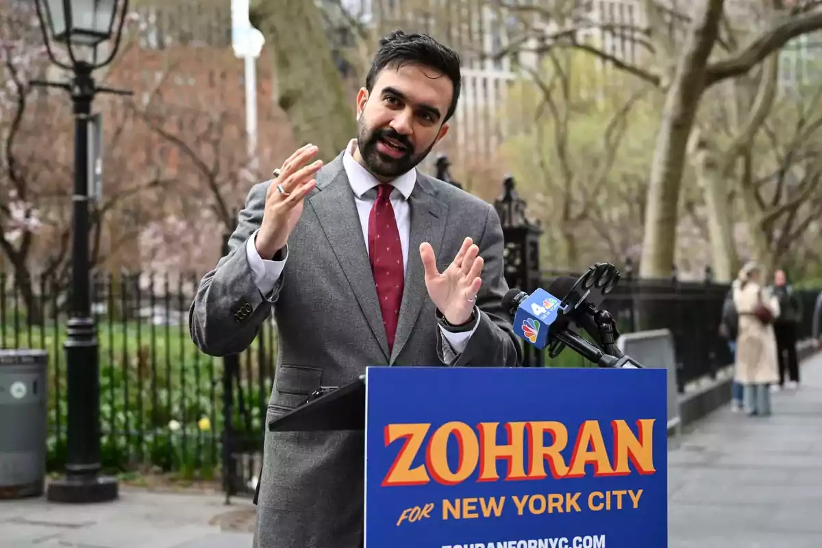 Hombre con barba y traje gris hablando en una conferencia de prensa al aire libre con un cartel azul que dice Zohran for New York City Hombre con barba y traje gris hablando en una conferencia de prensa al aire libre con un cartel azul que dice Zohran for New York City